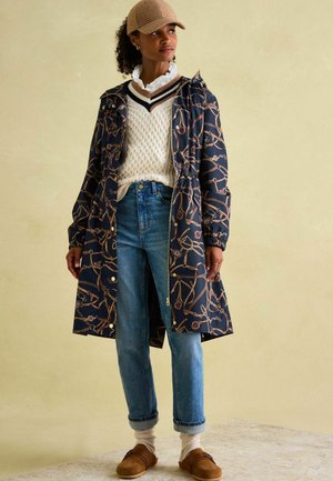 Marineblauer Mantel mit goldenem Kettendesign, gestrickter weißer Pullover, hellblaue Jeans, beige Slipper und eine hellbraune Mütze.