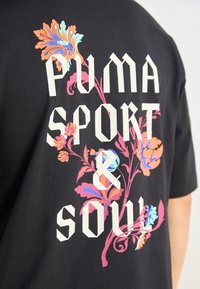 Svart t-shirt med fet vit text "PUMA SPORT & SOUL" och färgglada blommönster i rosa, blå och orange accenter. Mjuk tyg.