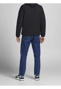 Svart puffjacka med horisontella sömmar, blå denimjeans och svarta och vita sneakers, sedd bakifrån.