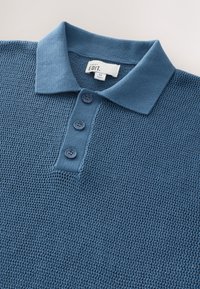 Gestricktes blaues Poloshirt mit strukturierter Webart, ausgestattet mit einem weichen Kragen und drei runden Knöpfen an der Vorderseite. Das Etikett trägt die Aufschrift "Next EDIT."