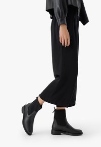 Czarne botki za kostkę wykonane z gładkiej skóry, z akcentami z ryżu dzianiny i uchwytem, zestawione z szerokimi czarnymi spodniami.