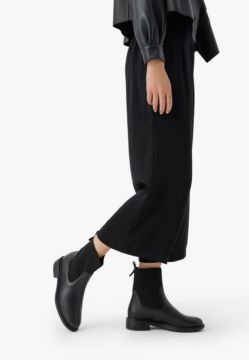 Czarne botki za kostkę wykonane z gładkiej skóry, z akcentami z ryżu dzianiny i uchwytem, zestawione z szerokimi czarnymi spodniami.