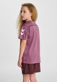 Fioletowy sportowy jersey z krótkim rękawem i białymi akcentami w kształcie chevronu; w połączeniu z ciemnobrązowymi szortami; wykonany z lekkiego materiału, idealny do aktywności fizycznej.