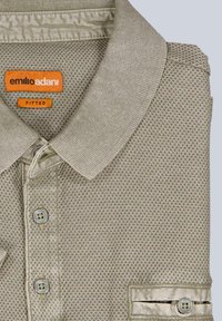 Figurbetontes Poloshirt in hellbeige, aus strukturiertem Stoff mit einem dezenten Muster. Verfügt über eine Knopfleiste, kurze Ärmel und ein oranges Markenlabel.