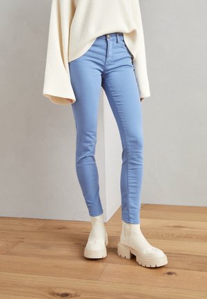 Femme portant un jean skinny bleu clair, un pull oversize crème et des bottines compensées épaisses ivoire, debout sur un sol en bois.