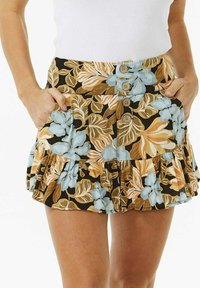 Frau in einem floral gemusterten Minirock mit blauen, beige und braunen Blättern, mit vorderem Knopfverschluss, Taschen und einem weißen ärmellosen Oberteil.