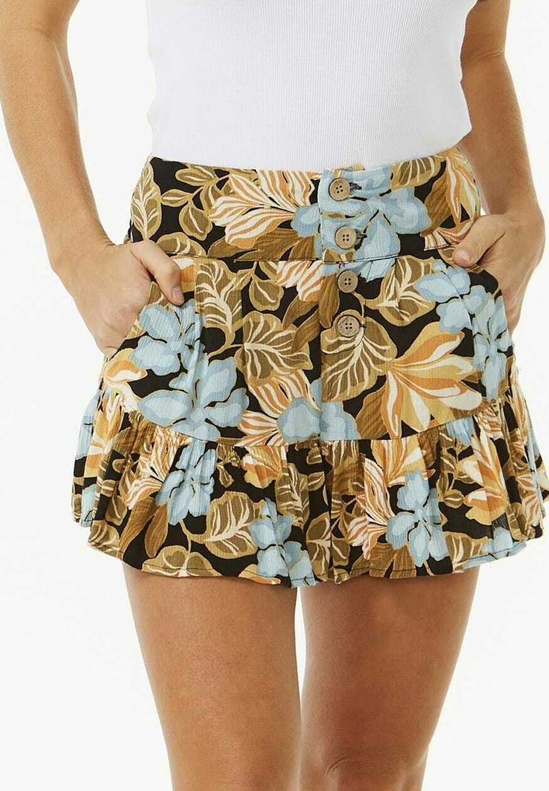 Frau in einem floral gemusterten Minirock mit blauen, beige und braunen Blättern, mit vorderem Knopfverschluss, Taschen und einem weißen ärmellosen Oberteil.