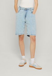 Person, der hellblaue Denim-Shorts bis zum Knie, weiße Socken und beige Sneakers mit blauen Streifen trägt, steht mit den Händen in den Taschen.