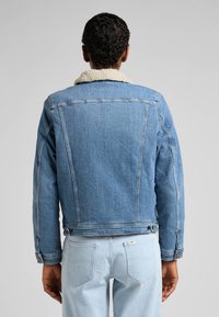 Džínová bunda ve světle modré barvě s bílým kožešinovým límcem, s kontrastním prošíváním a bočními kapsami, zobrazená zezadu.