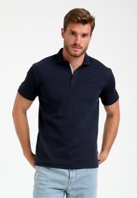 Polo majica temno moder barve, izdelana iz bombaža, z dvojnim gumbom, kratkimi rokavi in subtilnim logotipom na prsih. Nosi se z svetlimi jeans hlačami.