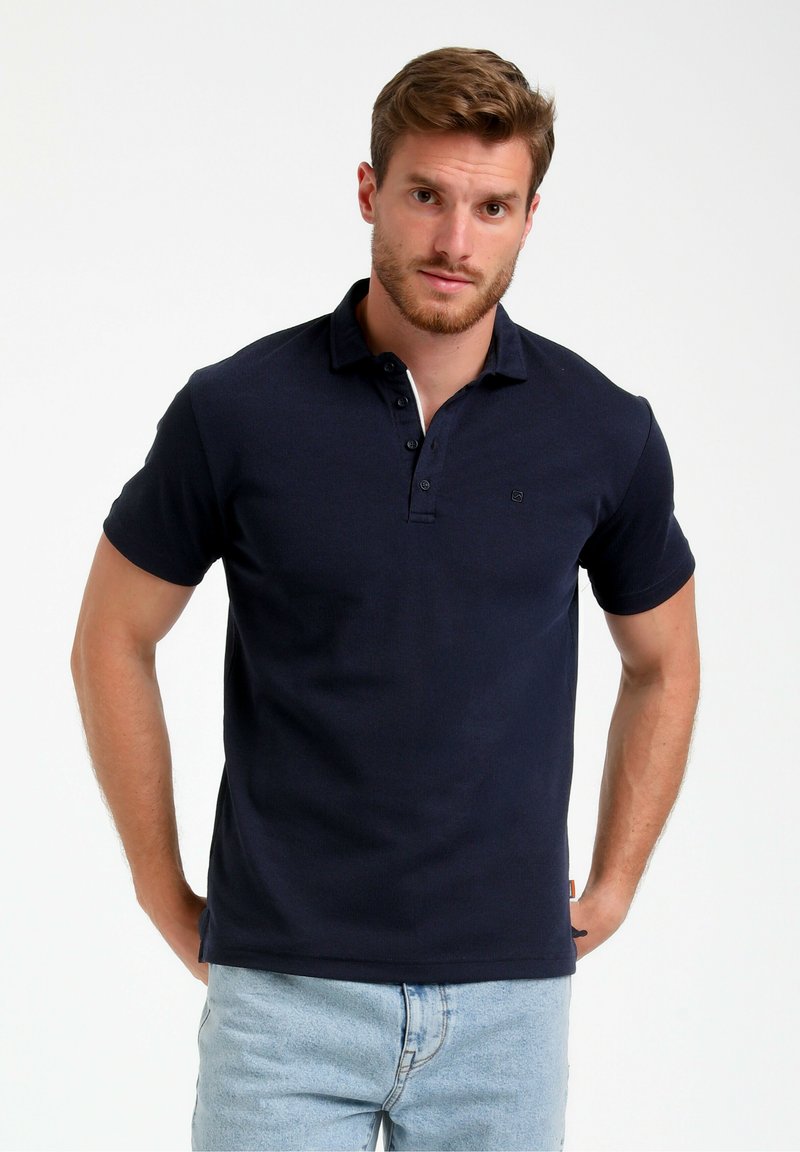 Polo majica temno moder barve, izdelana iz bombaža, z dvojnim gumbom, kratkimi rokavi in subtilnim logotipom na prsih. Nosi se z svetlimi jeans hlačami.