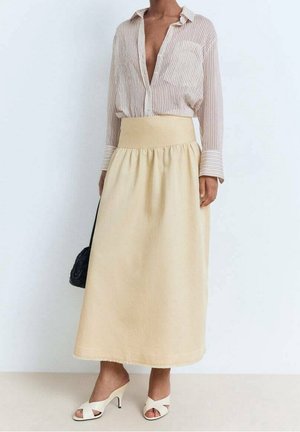 Kvinde iført en beige midi-nederdel, striped skjorte med åben krave, hvide hæle-sandaler og holder en sort håndtaske.