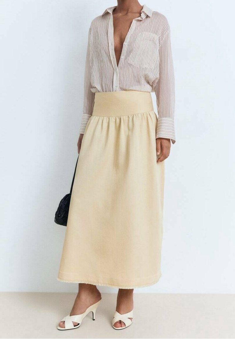 Femme portant une jupe midi beige, un chemisier rayé à col ouvert, des sandales blanches à talons et tenant un sac à main noir.