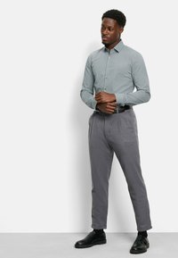 Button-up-Hemd in Hellgrün mit kleinem Muster, kombiniert mit grauen Anzughosen. Schwarze Slipper mit strukturierten Sohlen.