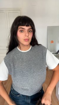 Sivi teksturiran brezrokavnik nad belo majico, v kombinaciji s temnoplavimi kavbojkami. V ozadju je ogledalo in oranžna svetilka.