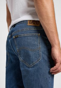 Jeans de denim azul con ligero desgaste, que presentan una etiqueta de cuero etiquetada "Lee" en la cintura, y costuras amarillas contrastantes a lo largo de los bolsillos.