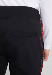 Schwarze Jogginghosen mit einem gerippten Bund, einer Gesäßtasche und roten vertikalen Details an der Seite. Hergestellt aus einem glatten Baumwollmischgewebe.