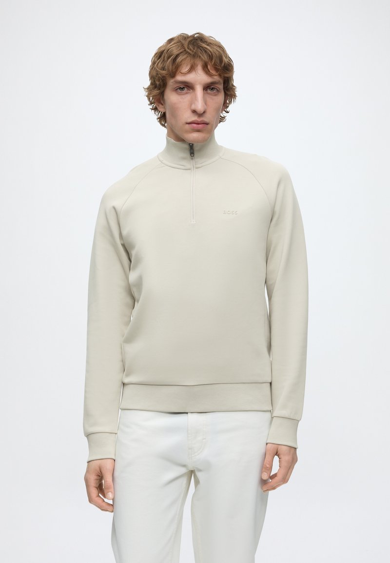 Jeune homme aux cheveux blonds bouclés portant un pull beige à demi-fermeture éclair et un pantalon blanc, debout devant un fond blanc uni.