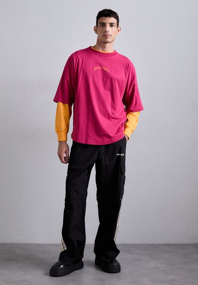 Rosa oversized t-shirt lagd over en gul langærmet skjorte, parret med sorte cargobukser med sidelærre og logo. Sorte sko.