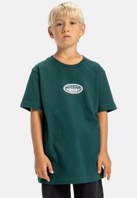 Camiseta de algodón verde oscuro con escote redondo, mangas cortas y un logo blanco central. La tela parece suave y ligera.