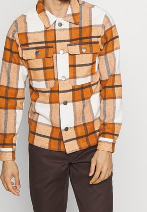 Mężczyzna ubrany w pomarańczową, białą i brązową koszulę w kratę zapinaną na guziki z kieszeniami z patkami na piersi oraz ciemnobrązowe spodnie na jednolitym tle.