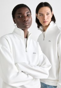 Sudaderas de forro polar blanco con media cremallera plateada y logotipo bordado, con un ajuste relajado y puños acanalados.