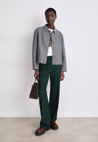 Casaco cinza em mistura de lã com botões dourados, t-shirt gráfica branca, calças largas verdes e sapatos castanhos de sola grossa. Pequena mala em bege com padrão de crocodilo.