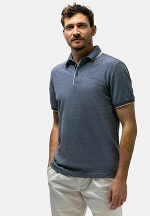 Man met kort bruin haar en baard, gekleed in een blauw poloshirt en lichtbeige broek, staand met één hand in de broekzak tegen een effen achtergrond.