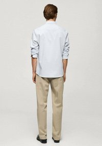 Hombre de pie de espaldas, vistiendo una camisa de manga larga azul claro con las mangas remangadas y pantalones beige, sobre un fondo liso.