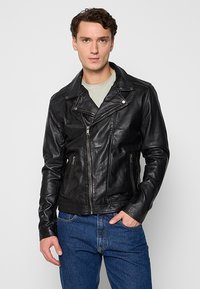 Veste de moto en cuir noir avec une fermeture éclair asymétrique, des revers larges et deux poches zippées à l'avant. Portée sur une chemise gris clair.