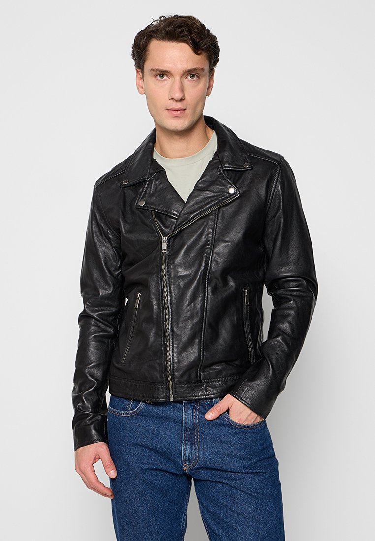 Veste de moto en cuir noir avec une fermeture éclair asymétrique, des revers larges et deux poches zippées à l'avant. Portée sur une chemise gris clair.