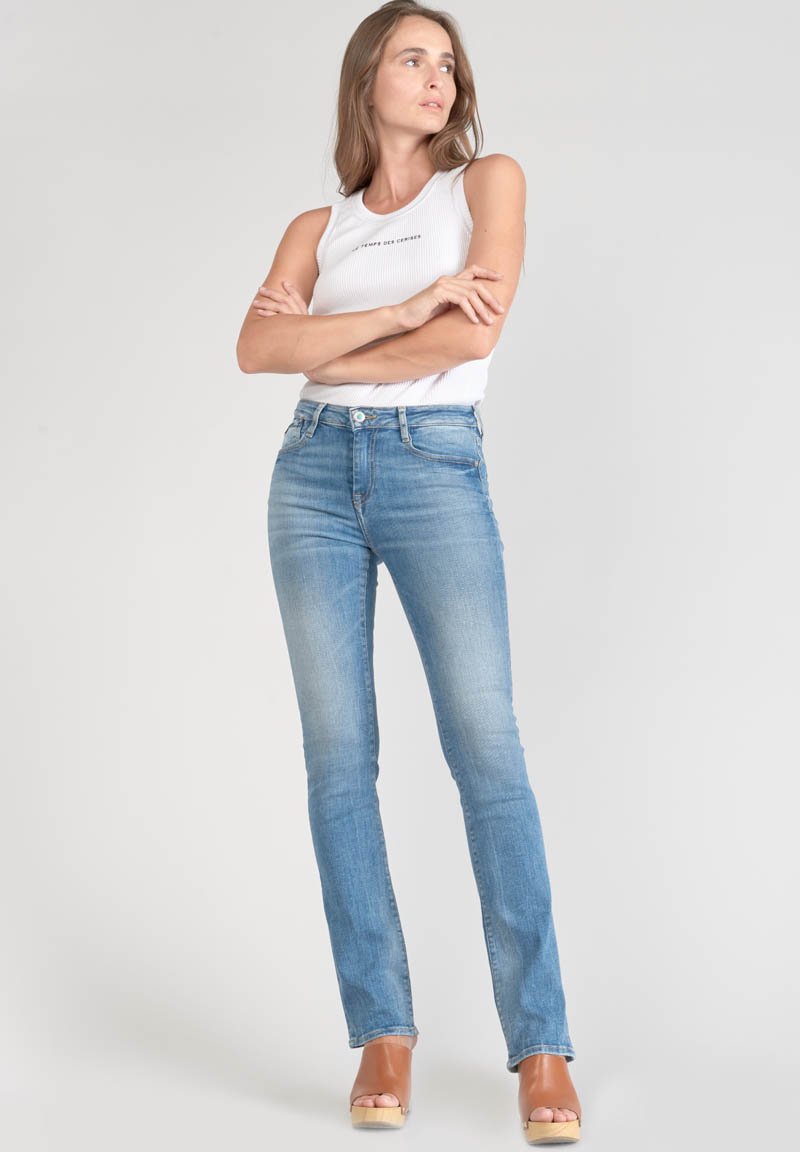 Jeans en denim bleu clair avec une coupe slim, un délavage usé, un design à cinq poches et une fermeture à bouton standard. Le modèle porte un débardeur blanc et des sandales à plateforme.