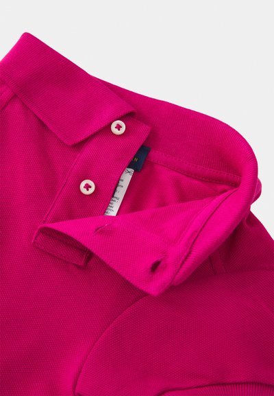 Cuello de polo rosa brillante con dos pequeños botones blancos, doblado para mostrar etiquetas internas y detalles de costura sobre un fondo liso.