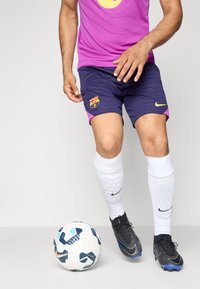 Violetinės ir tamsiai mėlynos futbolo šortai su „Barcelona“ logotipu, derinami su baltais kojiniais ir juodais sportbačiais, šalia raštuotas futbolo kamuolys.