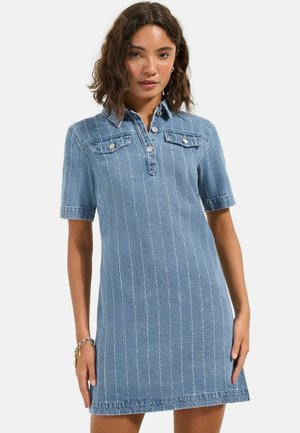 Žena oblečená v krátkym rukávom modrom rifľovom šatách s vertikálnymi švami, predným zapínaním na gombíky, golierom a náprsnými náprsné vreckami na bielom pozadí.