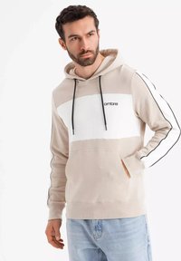 Béžová mikina s bílou přední částí, stahovací šňůrkou, bočními pruhy a klokankou. Obsahuje minimalistické logo na hrudi.
