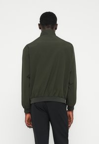 Άνδρας που φοράει μπουφάν bomber ελιάς και μαύρο παντελόνι, στέκεται γυρισμένος με την πλάτη προς το μέρος μας, σε απλό λευκό φόντο.