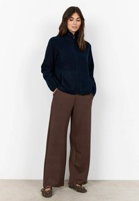 Vrouw in een marineblauwe fleecejack met rits, brede bruine broek en gestudeerde donkerbruine ballerina's staat voor een witte achtergrond.