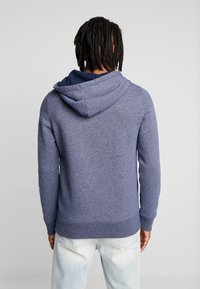 Person med dreadlocks som bär en blå hoodie, sedd bakifrån, stående framför en ljusgrå bakgrund, händerna vid sidorna.