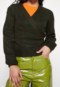 Dunkelgrüner, gestrickter Wickelpullover mit V-Ausschnitt und langen Ärmeln, darüber getragen ein leuchtend oranges Hemd. Kombiniert mit glänzenden grünen Hosen.