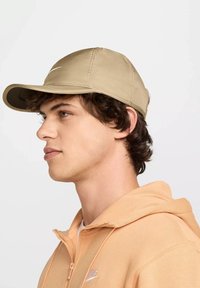  cuffia da baseball beige con visiera curva, cuciture a punto e logo sulla parte frontale. Indossata con un hoodie pesco, mostrata in vista di profilo.