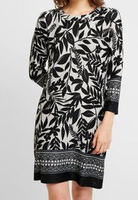 Robe à motifs floraux noirs et gris, manches longues, encolure ronde, avec détail décoratif à l'ourlet présentant des accents géométriques.