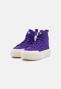 Purple high-top sneakers made of suede, featuring a thick white sole, white laces, and a round logo patch on the outer side.