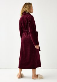 Burgundin kimono pehmeässä, pörröisessä kankaassa, jossa on huppukaulus, pitkät hihat ja soljettavalla vyöllä. Yhdistetty pehmeisiin beigeisiin tossuihin.
