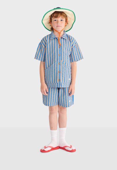 Enfant portant une chemise rayée bleue et marron clair avec un short, des chaussettes blanches et des tongs rouges, ainsi qu'un chapeau à large bord avec une lanière orange sous le menton.