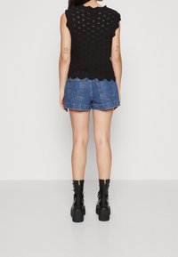 Černý pletený top s vlněnými okraji a vystřiženým vzorem, kombinovaný s modrými džíny šortkami. Modelka má na sobě černé platformové kotníkové boty.