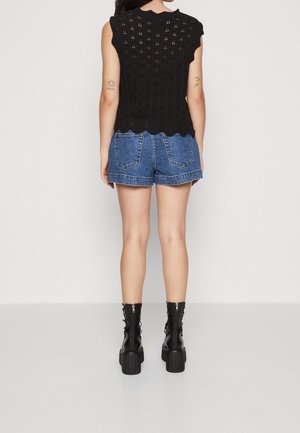 Czarny dzianinowy top z falbaniastymi brzegami i wycięciami, w połączeniu z niebieskimi szortami dżinsowymi. Modelka ma na sobie czarne botki na platformie.