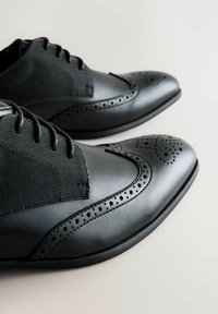 Pantofi de dress din piele neagră cu detalii perforate și panouri din țesătură texturată, cu șireturi, așezați pe o suprafață deschisă.