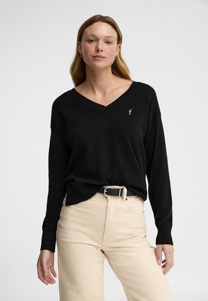 Kvinde med langt hår iført en sort V-hals sweater med en lille broderet logo og beige bukser med høj talje og sort bælte.