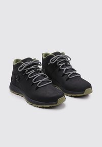 Zapatillas de cuero negro de media caña con cordones grises y acentos en verde oliva. Cuenta con un upper texturizado y suela de goma con un logo en el lateral.